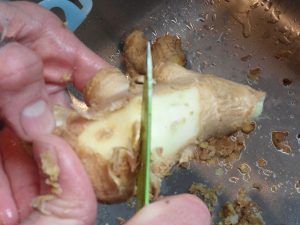A root of ginger is peeled with a knife, holding the knife perpendicular to the surface of the ginger.