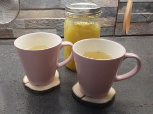 Two pink cups of ginger tea, a yellowish, slightly cloudy liquid. A mason jar full of the concentrated, yellowish ginger-sugar mixture is behind the cups.