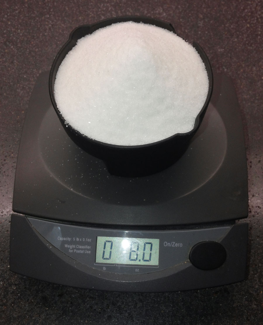 A cup of sugar is on a grey digital scale. The display shows 0 pounds, 8.0 ounces.
