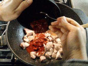 In a large black wok, red sauce is added over cooked pork. The pork is whitish in color.