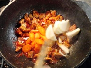 In a large black wok, carrots and onions are being added to stir-fried pork. The meat is red in color from the added sauce.