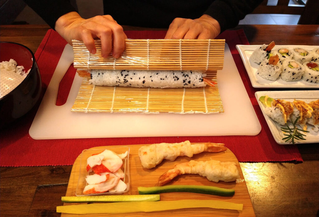 Tempura sushi roll in bamboo sushi roller-mat. Rolling process is finished. Inside ingredients are on a bamboo board below: crab meat, tempura shrimp, cucumber slices and pickled cucumber slice. 